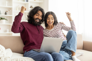 Giveaway, lottery, cashback. Excited happy beautiful indian couple using laptop, sitting on couch at home, gesturing and exclaiming, celebrating success, gambling on Internet, copy space