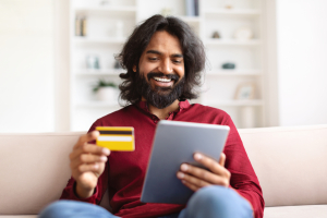 E-commerce, retail, financial app. Happy handsome young indian guy in smart casual banking or shopping from home, sitting on couch in living room, using digital tablet and credit card, copy space